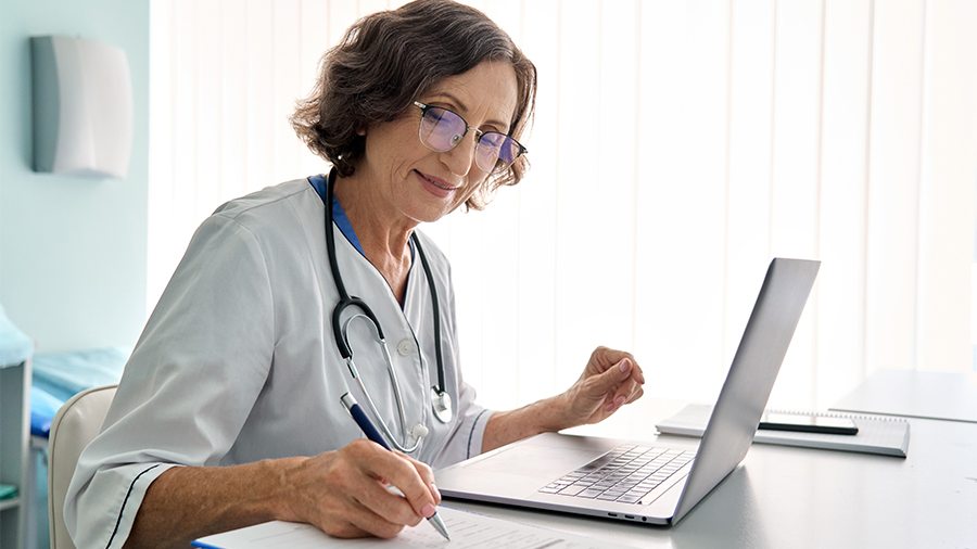 Physician sitting at computer and writing in note pad